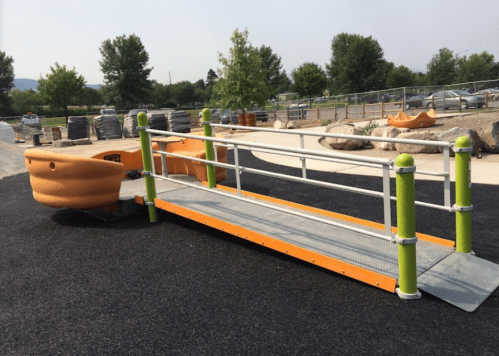 A colorful playground ramp with safety rails, leading to a play structure, surrounded by trees and gravel.